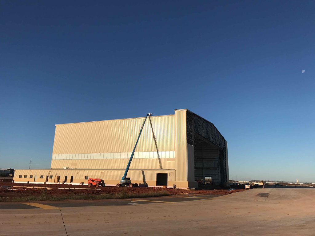 A large beige airport hangar under construction, with a boom lift and workers near the roof—showcasing impressive civil engineering. The sky is clear and blue, and the open tarmac stretches out in front of the building.