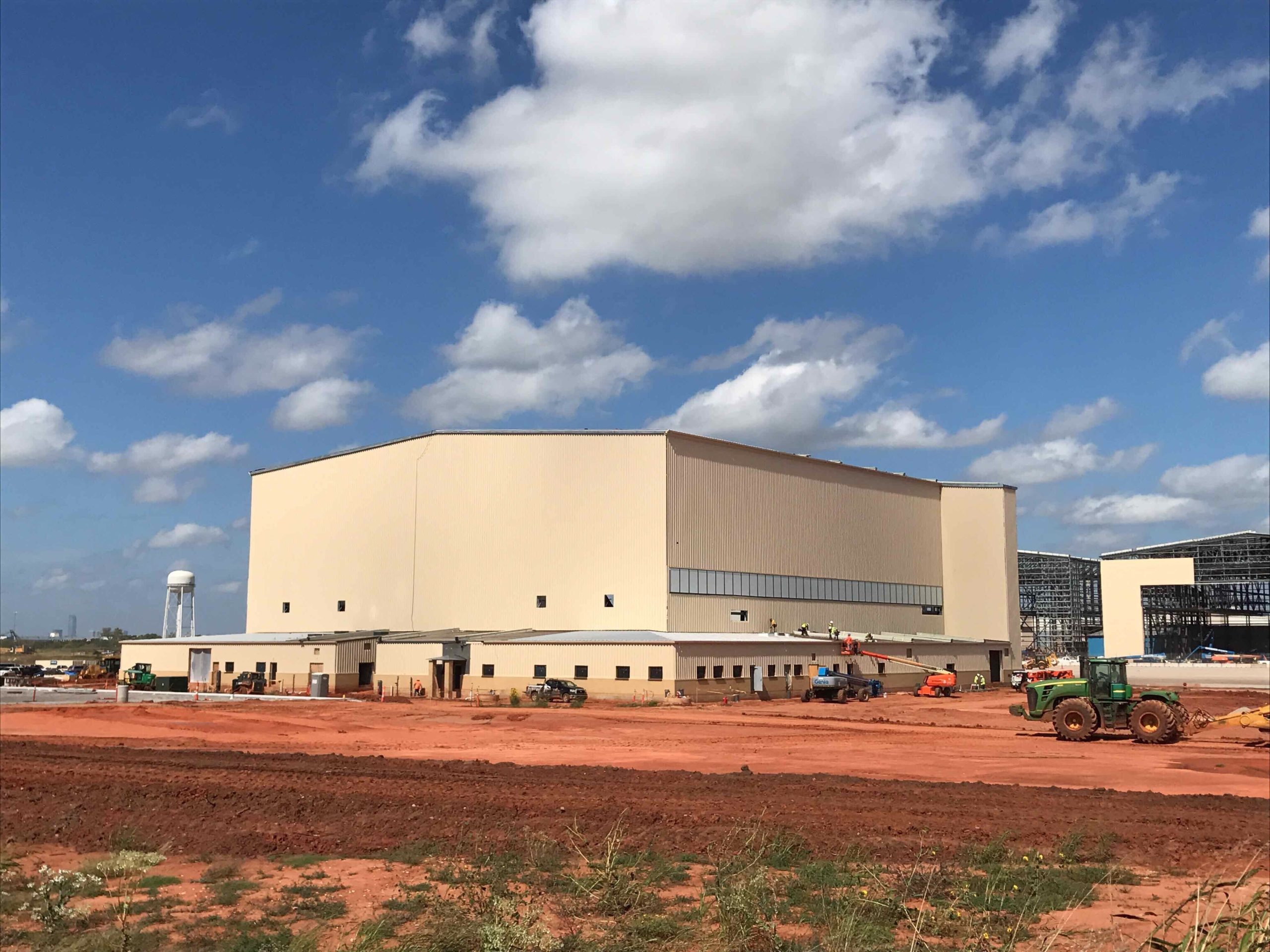 A large beige industrial building under construction stands on a red dirt lot, with Colorado engineering vehicles and equipment nearby under a partly cloudy blue sky.