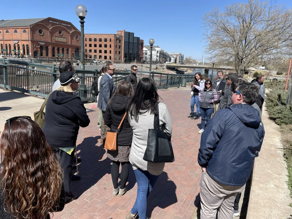 A group of people stand on a brick walkway outside, listening to a structural engineer speak. Trees without leaves and large brick buildings are visible in the background under a clear blue sky.
