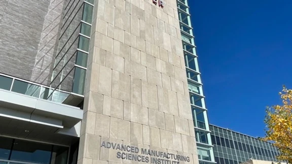 A tall modern building with large windows and tan stone walls displays the words Advanced Manufacturing Sciences Institute near the entrance, highlighting expertise in civil engineering, under a clear blue sky.