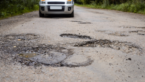 A white car approaches on a rural road filled with numerous large potholes, surrounded by greenery—perhaps a challenge fit for a skilled Colorado engineering team.