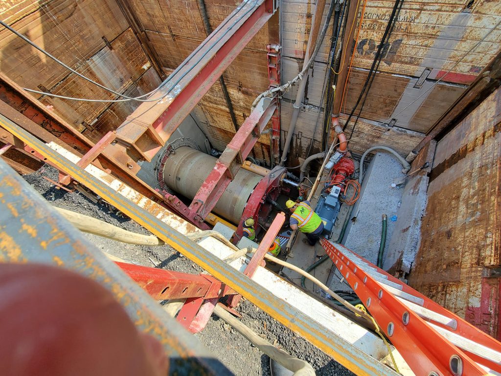 A construction worker in a safety vest and helmet operates machinery inside a deep, narrow excavation site with steel beams, pipes, cables, and wooden retaining walls under the supervision of a structural engineer.