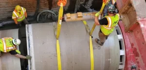 Three construction workers in safety gear guide large concrete pipe sections into place inside a trench, using yellow lifting straps attached to heavy machinery above—a skilled display of civil engineering expertise.