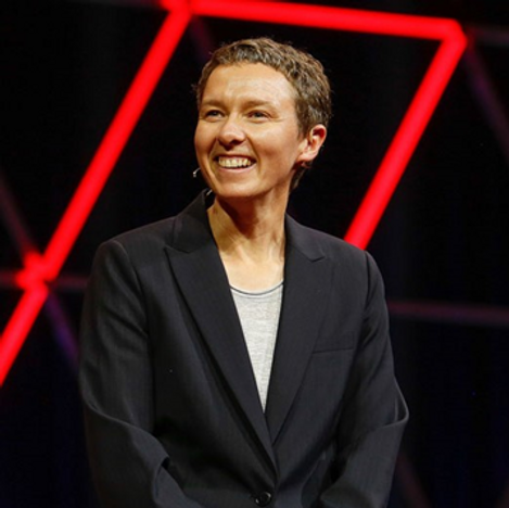 A person with short brown hair smiles while wearing a dark blazer and light shirt, standing on stage with red geometric light shapes in the background, embodying the innovation seen in colorado engineering.