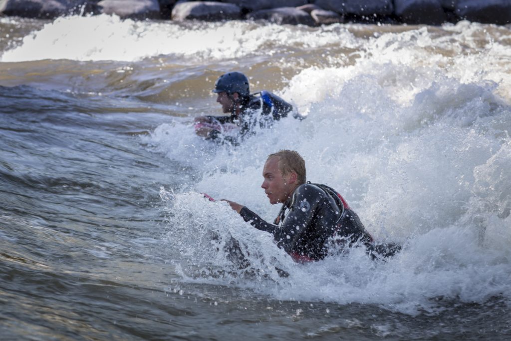 Two people wearing wetsuits and helmets ride waves on bodyboards in a river, surrounded by splashing water, showcasing the thrill of whitewater engineer adventures with rocks visible in the background.