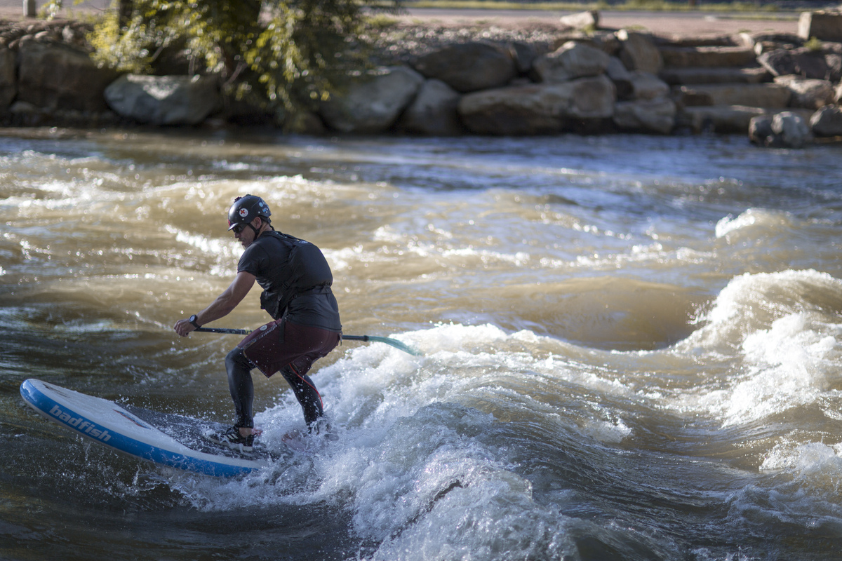 A person wearing a helmet and wetsuit rides a blue paddleboard on a river with whitewater rapids, showcasing the adventure often designed by a skilled whitewater engineer; rocks and trees line the riverbank in the background.