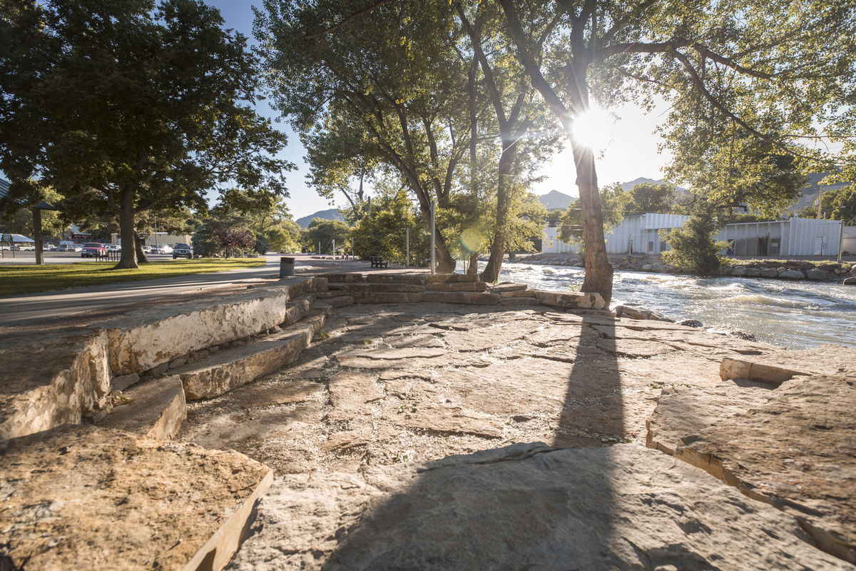 Sunlight filters through leafy trees onto stone steps by a flowing river. A Colorado engineering firm designed the area, with buildings and parked cars in the background and a well-maintained green lawn lining the walkway alongside the water.