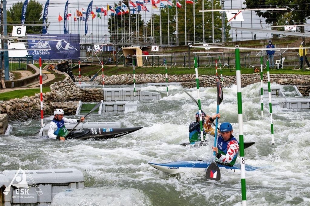 Three athletes in kayaks paddle through whitewater rapids on a slalom course, designed by a skilled whitewater engineer. Numbered gates span the rushing water as officials and spectators watch from the background, with flags waving overhead.
