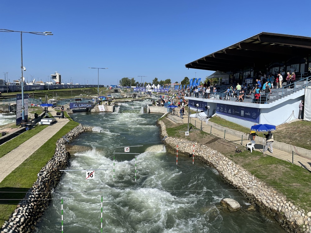 A whitewater slalom course, expertly designed by a whitewater engineer, features rapid water, gates, and numbered poles. Spectators watch from a shaded grandstand on the right under a clear blue sky.