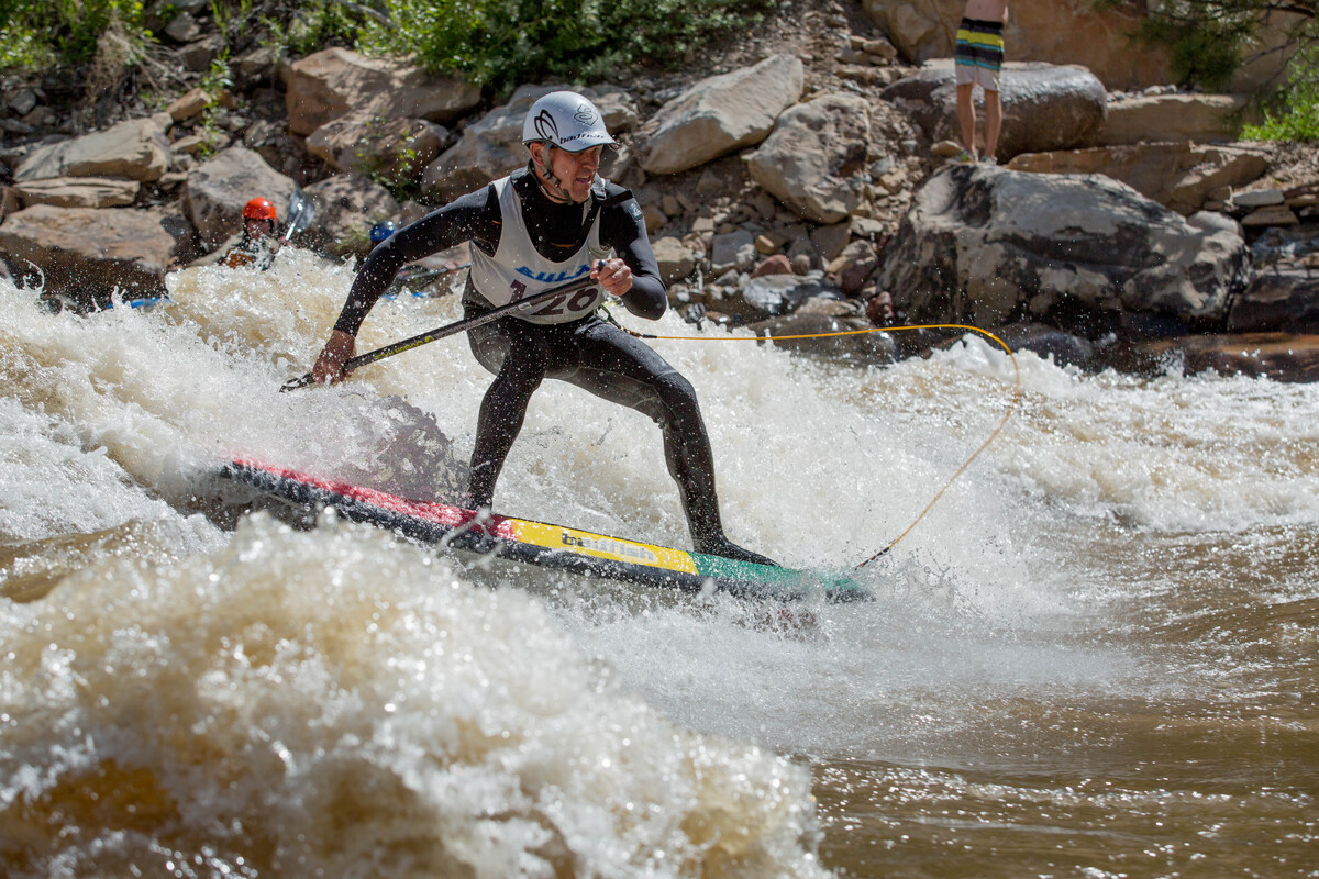A person in a helmet and wetsuit competes in a river paddleboarding race, balancing amid whitewater rapids—a thrilling scene that might inspire any Denver engineer or civil engineering enthusiast standing on the rocky shore.