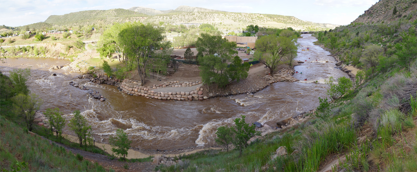 Panoramic view of a river with rapids, designed by a skilled whitewater engineer, flowing through a green landscape with trees by the shore, park pathways, rocks, hills, and a blue sky in the background.