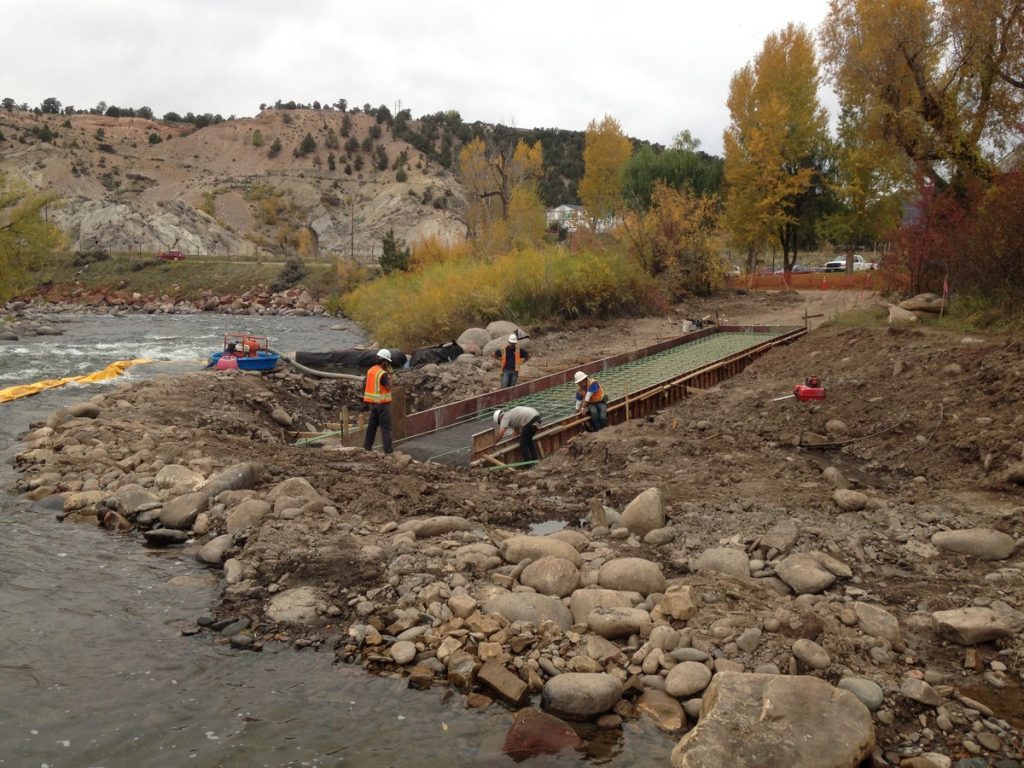 Construction workers in safety vests, guided by expert civil engineering, build a concrete structure by a riverbank, surrounded by rocks and autumn trees, with hills and a cloudy sky in the background.