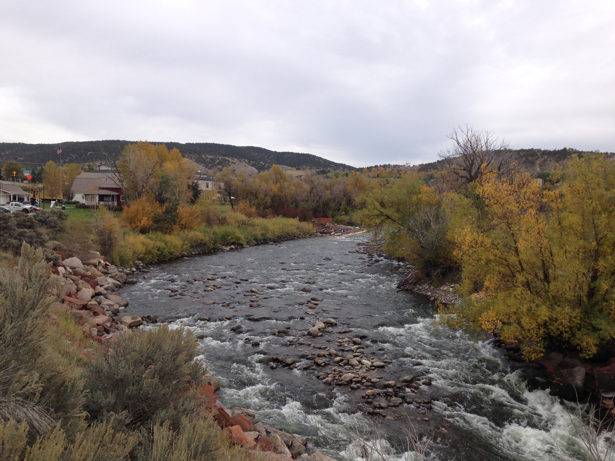A river flows through a rocky, tree-lined landscape with autumn foliage. Houses and hills designed by a structural engineer are visible in the background under a cloudy sky.