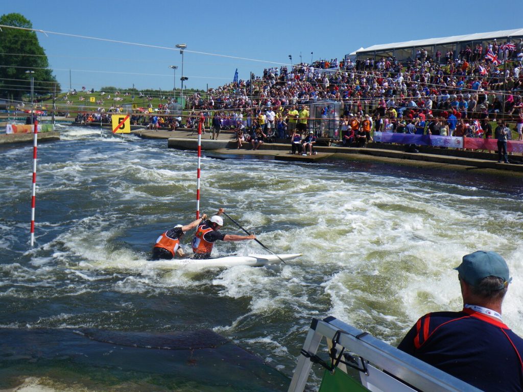 Two athletes in orange vests paddle a canoe through whitewater rapids on a slalom course designed by Colorado engineering experts, with a large crowd watching from the stands and a judge observing from the foreground.