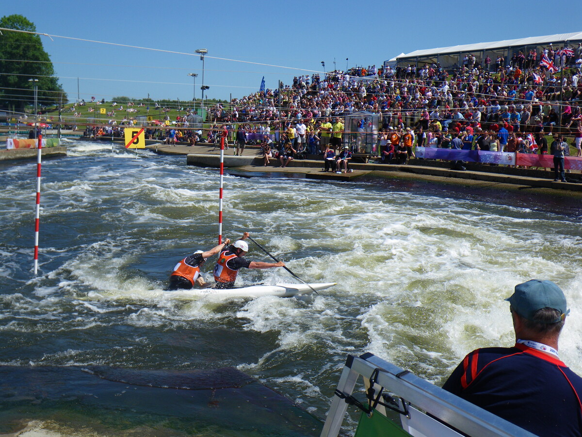Two athletes in orange vests paddle a canoe through whitewater rapids on a slalom course designed by Colorado engineering experts, with a large crowd watching from the stands and a judge observing from the foreground.