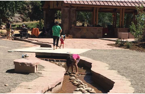 A young girl bends over to play in a shallow stream bordered by rocks, possibly designed by a whitewater engineer, while two other people walk nearby toward a shaded pavilion in a park-like setting.