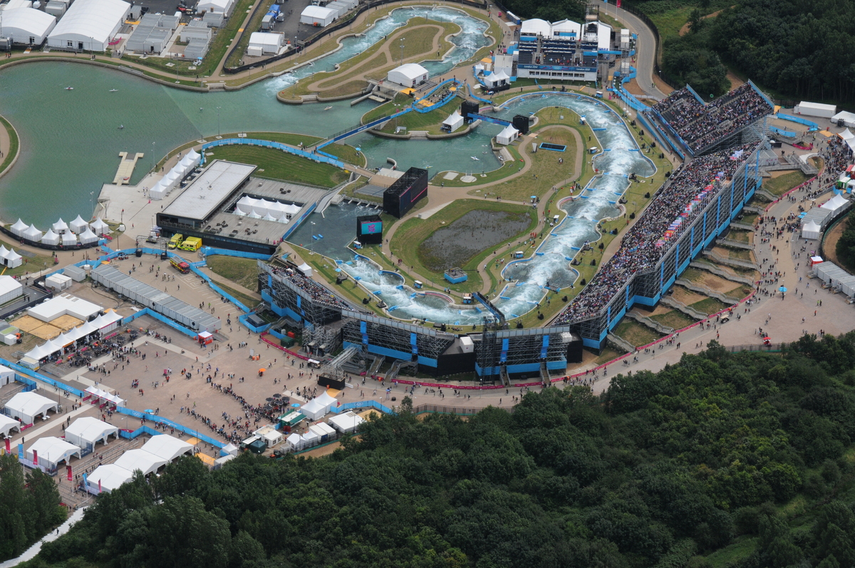 Aerial view of an outdoor whitewater rafting or kayaking venue designed by a whitewater engineer, with winding artificial river, stadium seating filled with spectators, surrounding tents, and crowds walking along pathways.