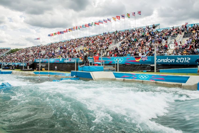 A large crowd fills stadium stands decorated with international flags at the London 2012 Olympics, as a whitewater engineers expertise is showcased in the turbulent course flowing in the foreground.
