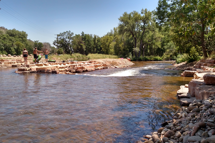 Three people stand on rocky edges by a shallow river under a clear blue sky, with trees and grassy banks in the background—a peaceful scene that might inspire any civil engineering or structural engineer in Denver. Smooth stones line the shoreline.