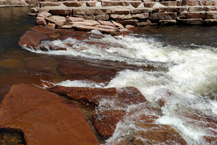 Fast-moving water flows over reddish-brown rocks, bordered by a wall of stacked light-colored stones—an impressive showcase of civil engineering—creating a small, foamy rapid in either a natural or man-made setting.