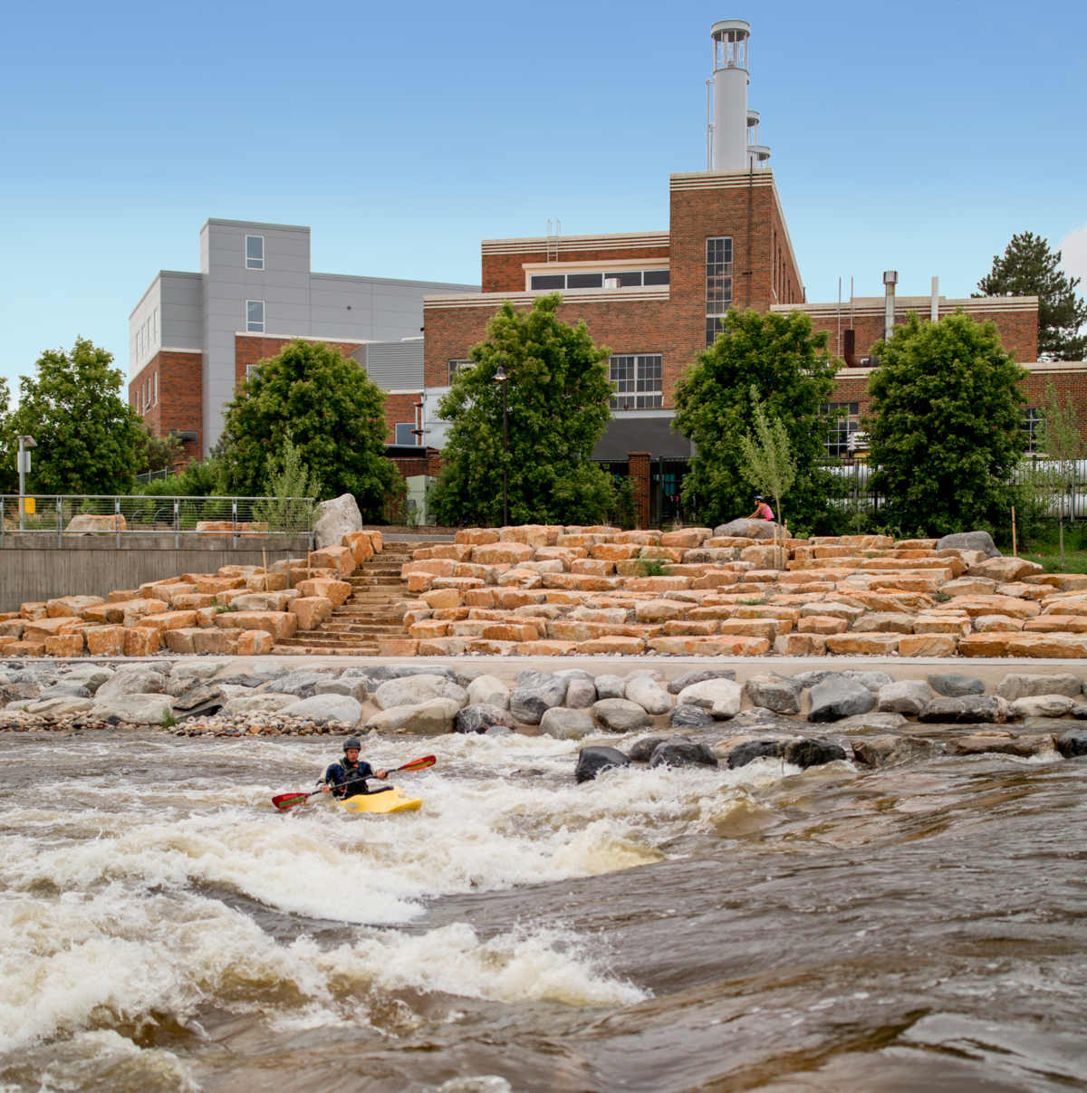 A person kayaks through whitewater rapids in a river designed by a skilled whitewater engineer, with tiered stone steps and a large brick building with a smokestack and trees in the background. Another person sits on the rocks near the riverbank.