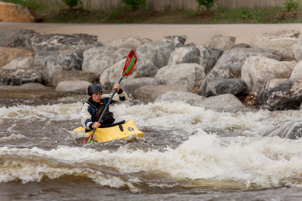 A person wearing a helmet and life jacket paddles a yellow kayak through choppy whitewater near large rocks on a riverbank, showcasing the challenge civil engineering faces managing river flow and safety.