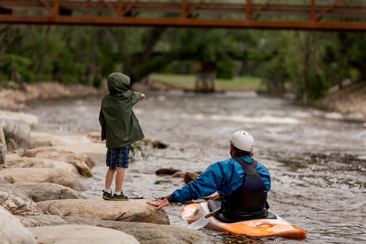 A child in a green jacket stands on rocks by a river, pointing ahead, while an adult in a helmet and blue jacket sits in a kayak in the water. A bridge—perhaps designed by a civil engineer—spans the background amid leafy trees.