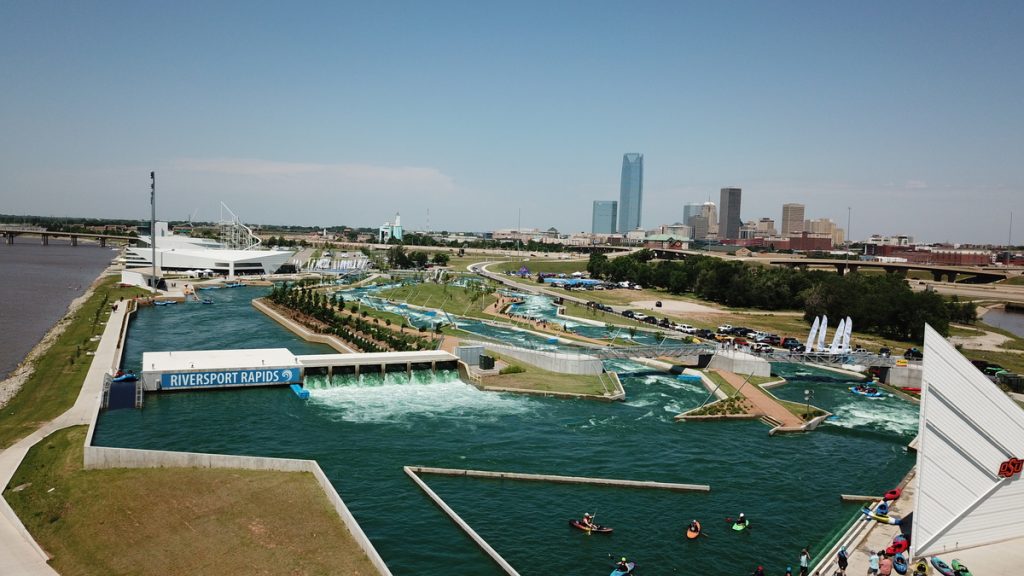 Aerial view of Riversport Rapids whitewater center in Oklahoma City, showcasing winding water channels crafted by expert whitewater engineers, with adjacent river, people in rafts, modern buildings, and the city skyline under a clear sky.