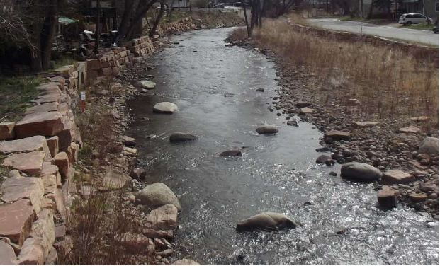 A gently flowing creek with large rocks along its shallow bed, bordered by stone walls that reflect careful civil engineering. Trees and a road run parallel to the creek on the right side of the image.