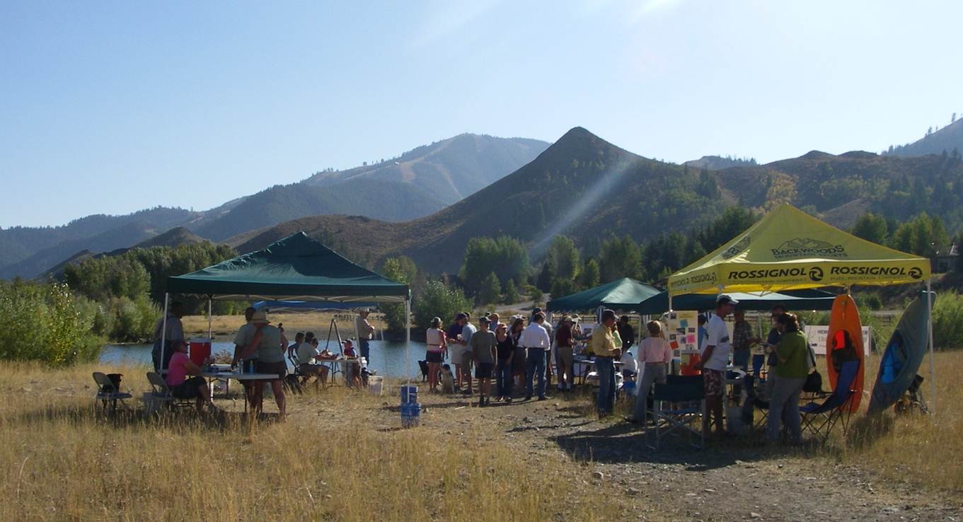 A group of people gather under tents by a lake in a grassy field, with mountains and trees in the background, on a sunny day. Tables with displays and activities are set up for an outdoor event featuring colorado engineering projects.