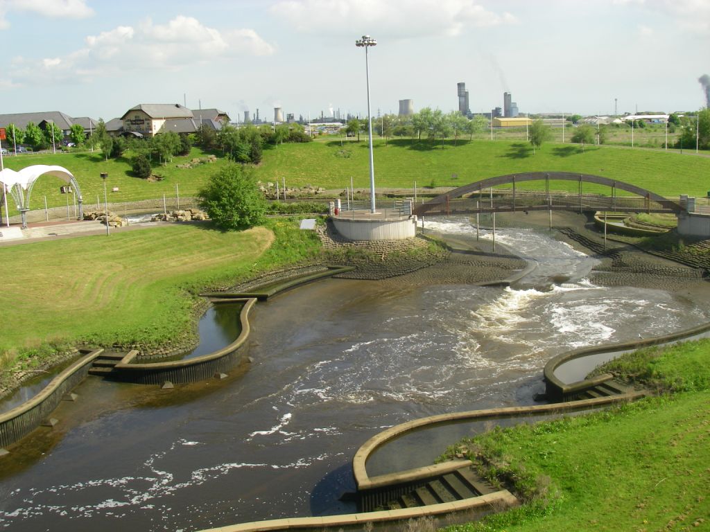 A landscaped urban park designed by a Denver engineer features a winding artificial watercourse with small rapids, green lawns, a footbridge, and buildings in the background under a partly cloudy sky.
