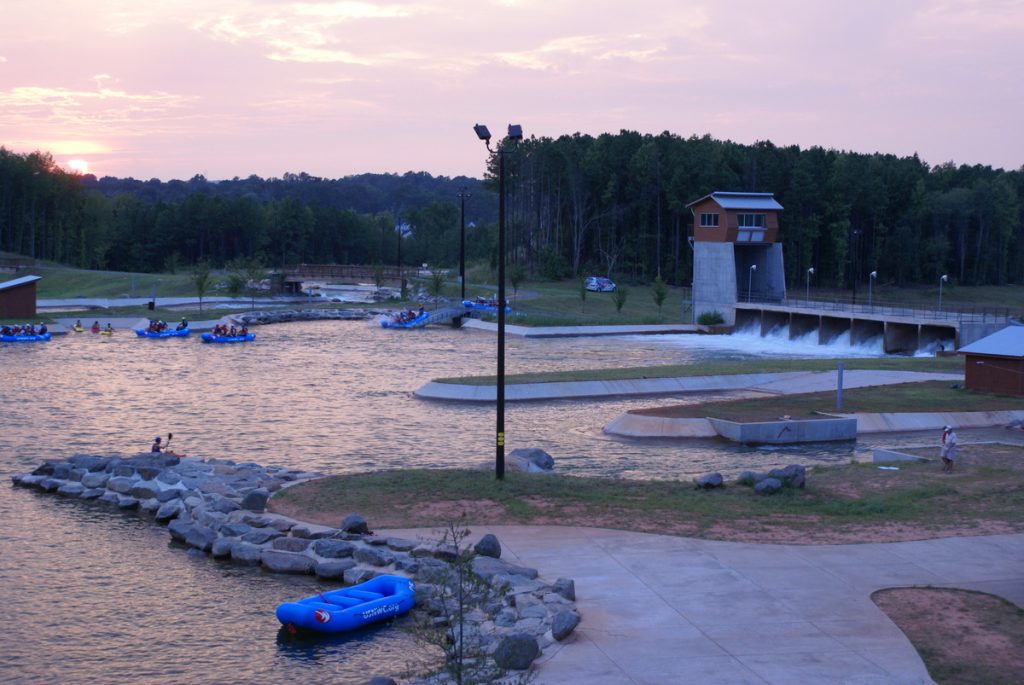 People are rafting on a man-made whitewater course at sunset, surrounded by trees and grassy areas. Designed with civil engineering expertise, the scene features concrete structures, nearby buildings, and an empty blue raft resting on the shore.