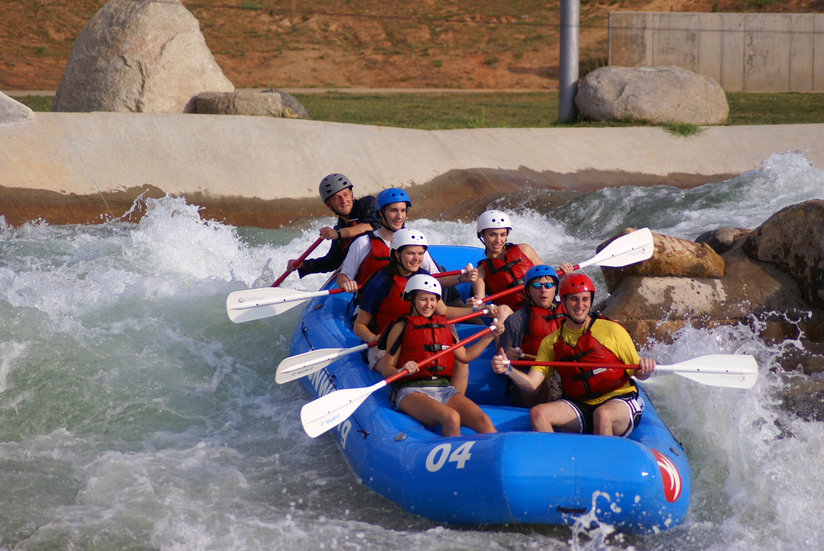 A group of eight people wearing helmets and life jackets paddle a blue inflatable raft through whitewater rapids, surrounded by rocks and choppy water—perhaps unwinding after a week as Colorado engineering professionals.