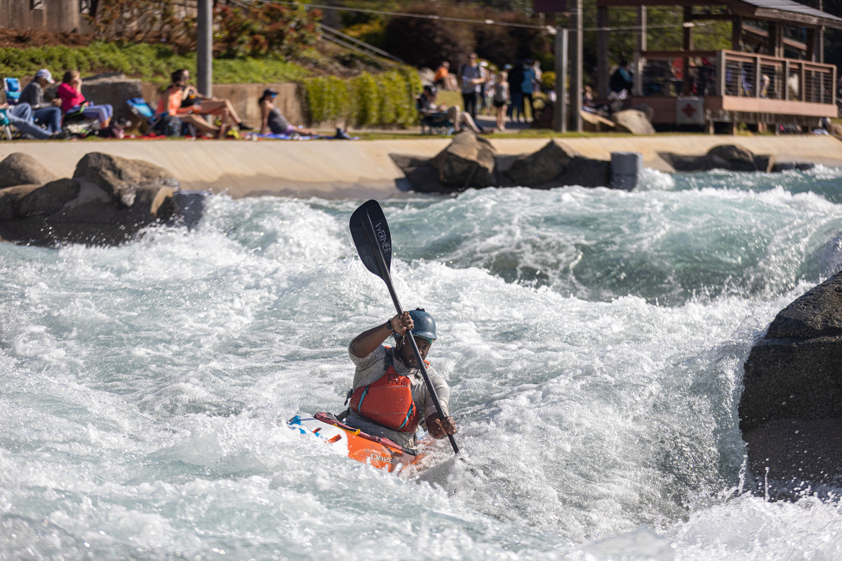 A kayaker wearing a helmet and life jacket paddles through rough whitewater rapids, while spectators—including a Denver engineer—watch from the riverbank in the background.