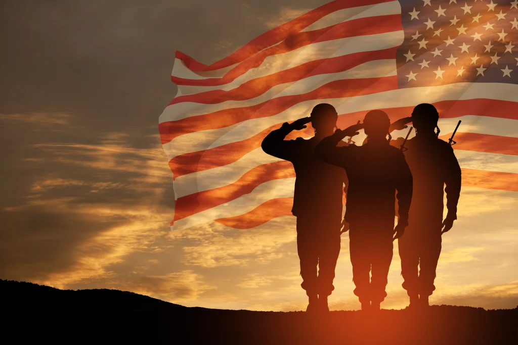 Three soldiers stand in silhouette, saluting at sunset with a large American flag in the background, symbolizing patriotism, military service, and the dedication of SDVOSB engineering professionals.