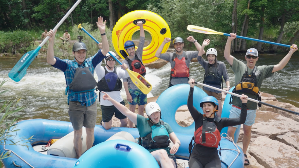 A group of nine people, including a Denver engineer, wearing helmets and life vests pose excitedly with paddles and inflatable tubes by a river, surrounded by trees and water, celebrating an outdoor rafting adventure.