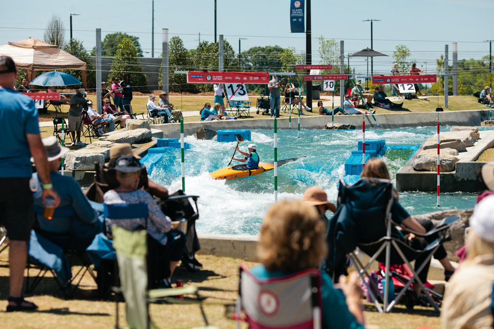A kayaker paddles through a whitewater course designed by a skilled whitewater engineer as people sit in chairs watching from the grassy bank. Numbered gates hang above the water, and spectators are enjoying the sunny outdoor event.