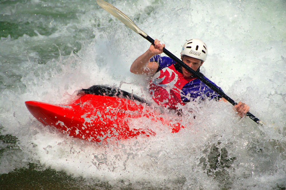 A kayaker wearing a helmet and colorful shirt paddles through rough, splashing whitewater in a bright red kayak—channeling the spirit of a whitewater engineer amid foamy waves.