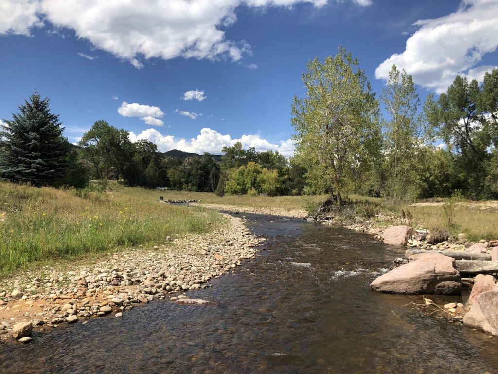 A shallow stream flows through a grassy, rocky landscape bordered by trees under a blue sky with scattered clouds—an inspiring scene for any civil engineering or structural engineer seeking nature’s harmony as sunlight highlights the greenery and stones by the water’s edge.