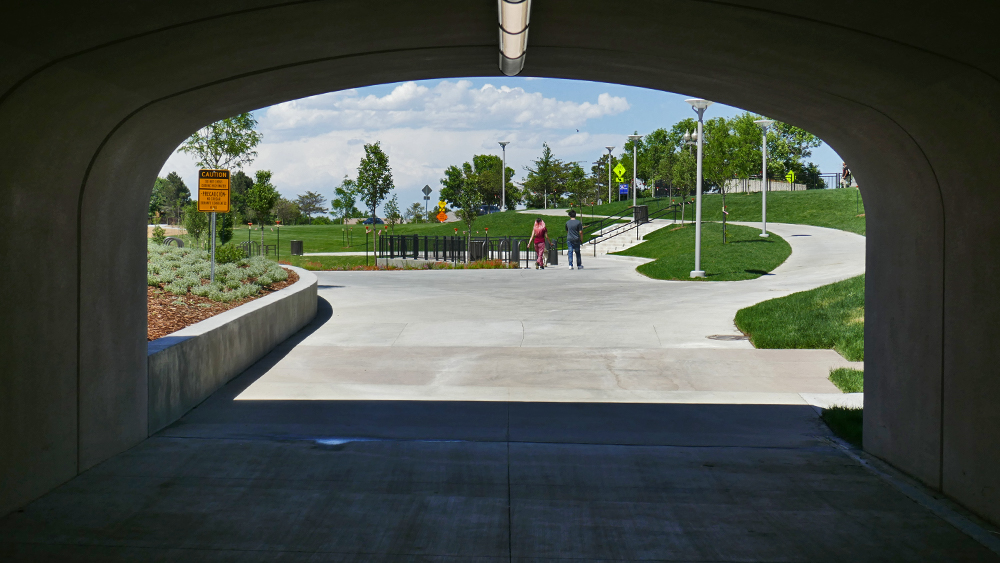 View from inside a tunnel designed by a structural engineer, looking out onto a sunny park with green grass, trees, a sidewalk, and three people walking away in the distance. A yellow sign is visible on the left side.