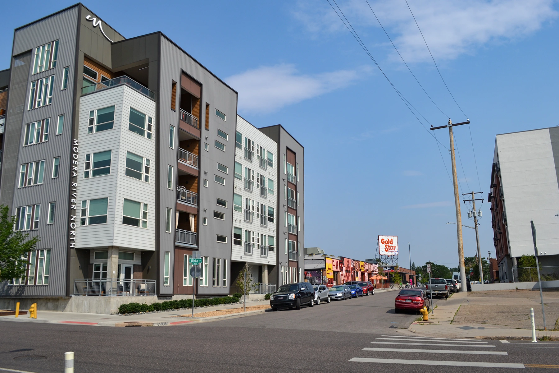 A modern apartment building stands on a quiet urban street lined with parked cars. In the distance, small colorful businesses—including a nod to Colorado engineering—join the “Gold Medal Bar” sign under a clear blue sky.
