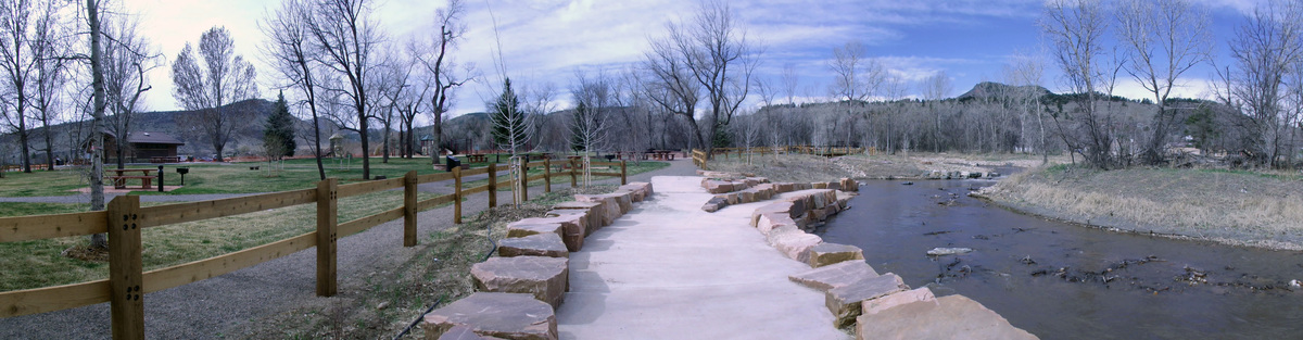 A paved path bordered by large rocks runs alongside a creek in a park with leafless trees, grassy areas, and picnic tables—an inviting scene even a Denver engineer or structural engineer might admire beneath the partly cloudy sky and distant hills.