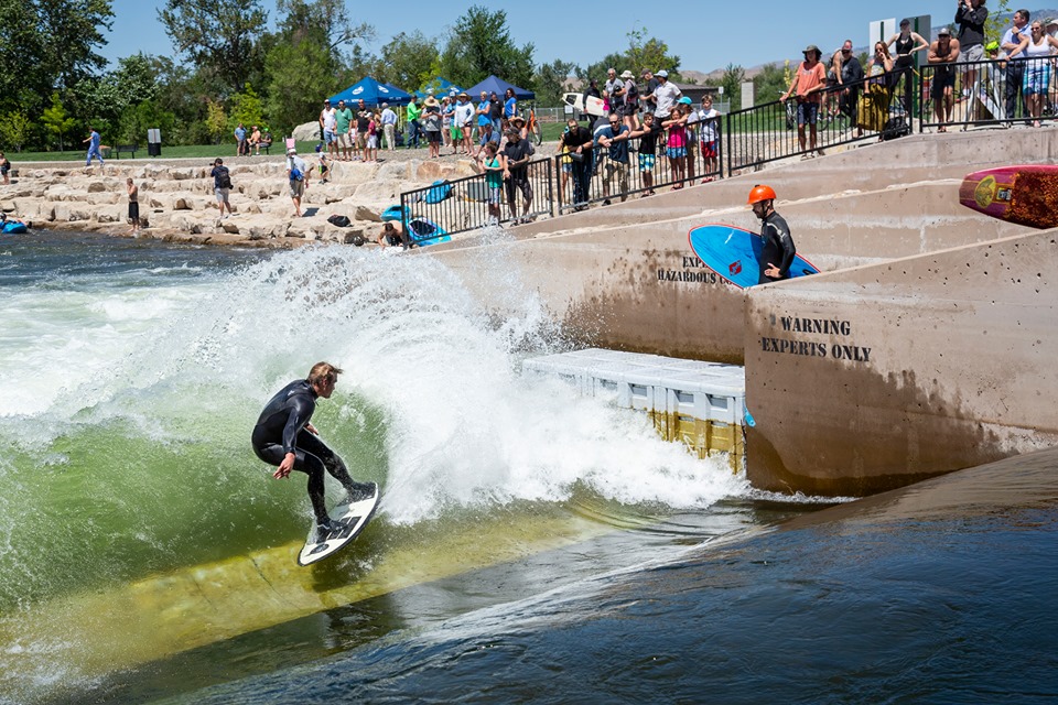A surfer in a wetsuit rides an artificial wave at a river park, designed by a whitewater engineer, as spectators watch from behind a fence. Another person with a helmet and surfboard waits nearby on a concrete platform.