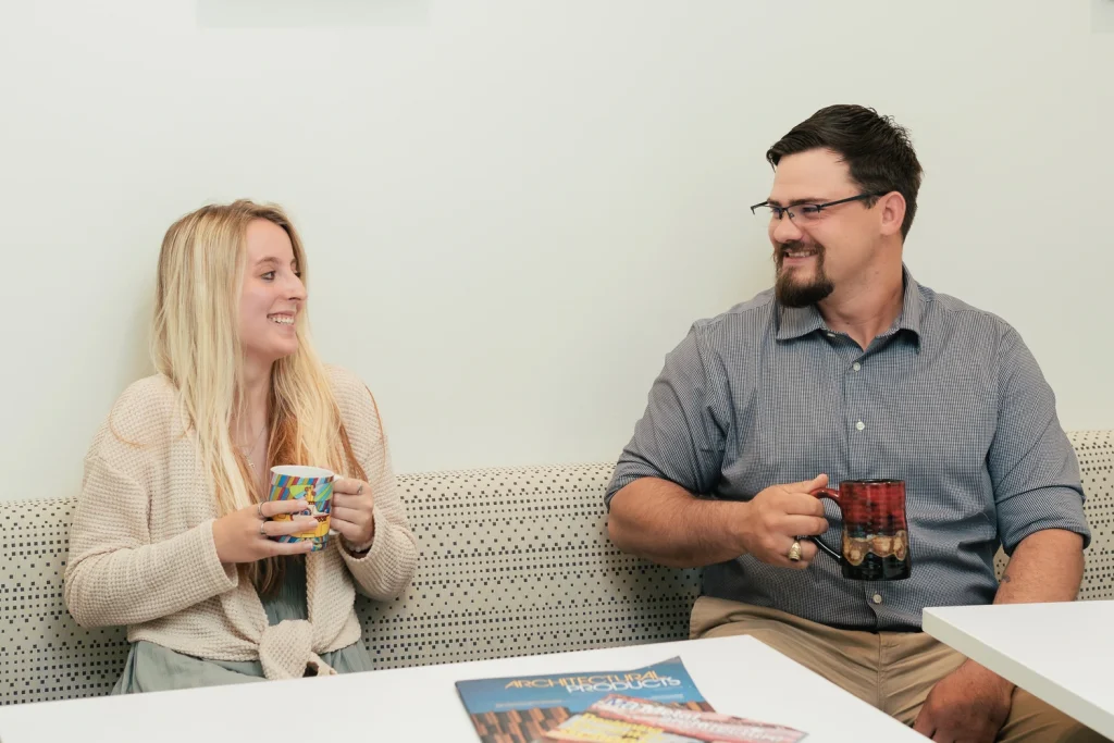 A woman and a man sit on a bench, smiling and holding mugs, enjoying a friendly conversation about Colorado engineering. A magazine and snack bars are on the table in front of them.