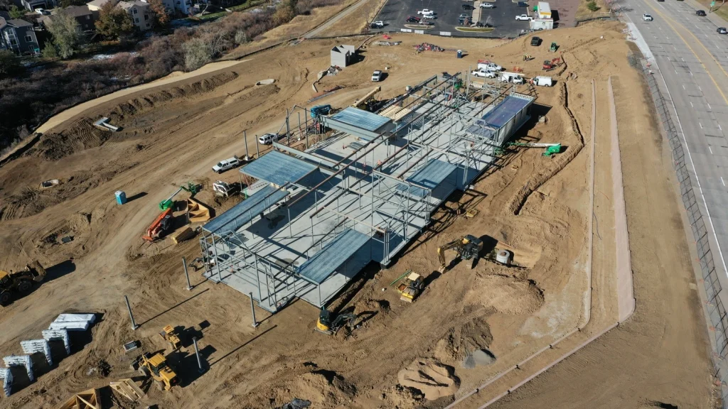 Aerial view of a construction site with a large building framework, construction vehicles, equipment, and dirt piles—showcasing the work of Colorado engineering experts; cars are parked nearby and a road runs along the right side of the image.