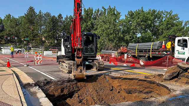 A construction excavator is digging a large hole in a city street, surrounded by traffic cones, barriers, construction materials, and workers. A structural engineer oversees the site, with trees and construction vehicles visible in the background.