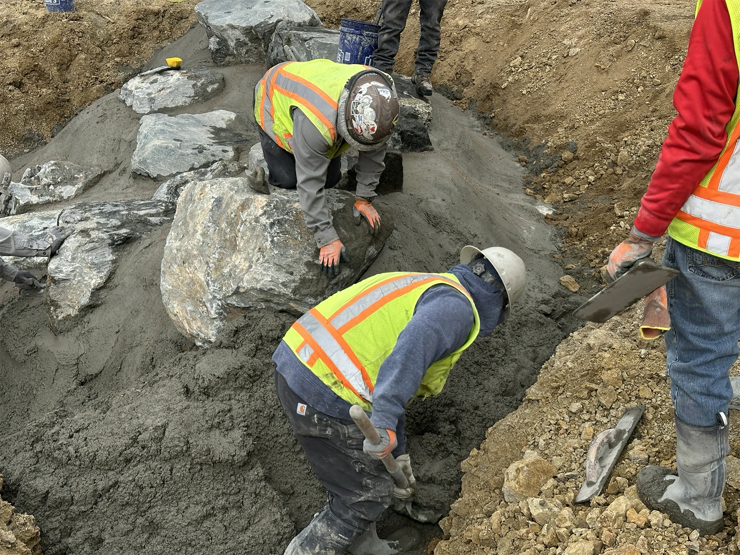 Three construction workers in safety vests and helmets, part of a Colorado engineering team, work together placing large rocks and spreading cement in a trench at a construction site surrounded by dirt piles.