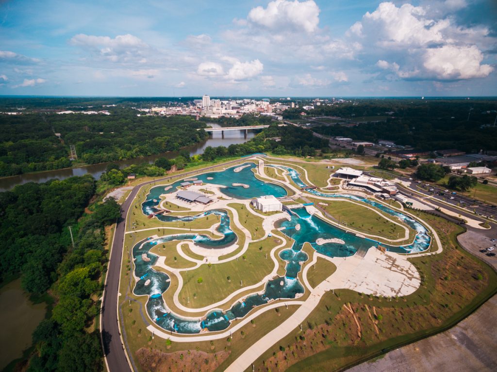 Aerial view of a large, winding lazy river water park designed with expert civil engineering, surrounded by greenery. City skyline and scattered clouds are visible in the distance, with multiple pools and walking paths integrated into the landscape.
