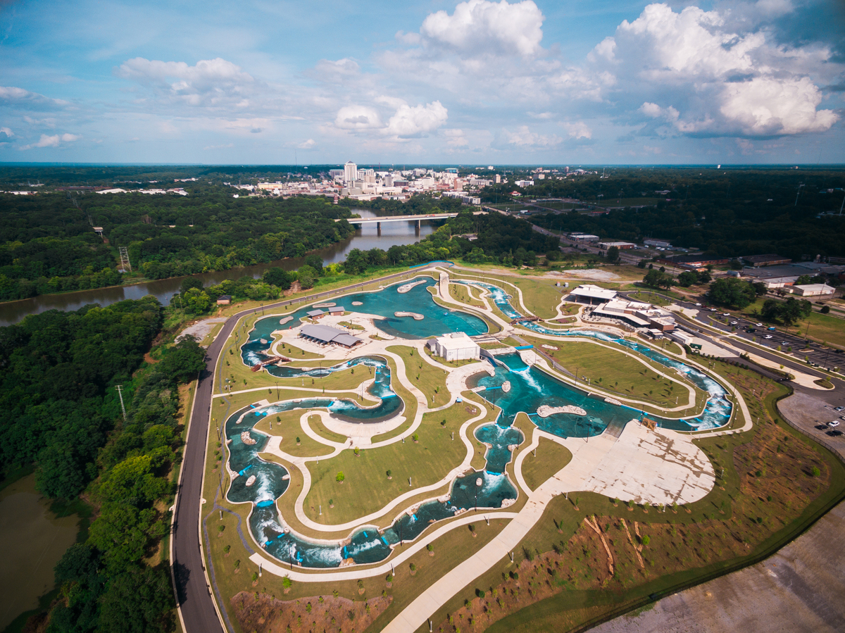 Aerial view of a large, winding lazy river water park designed with expert civil engineering, surrounded by greenery. City skyline and scattered clouds are visible in the distance, with multiple pools and walking paths integrated into the landscape.