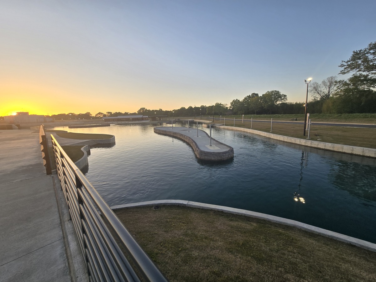 A winding man-made water canal, expertly designed by civil engineering professionals, features walkways and lampposts at sunset, bordered by grassy areas and trees under a clear sky.